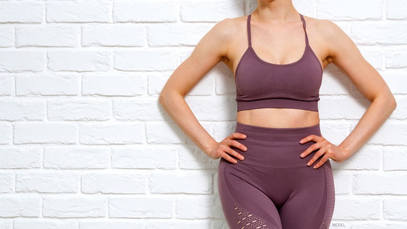 A woman in sports bra and yoga pants leaning against brick wall.