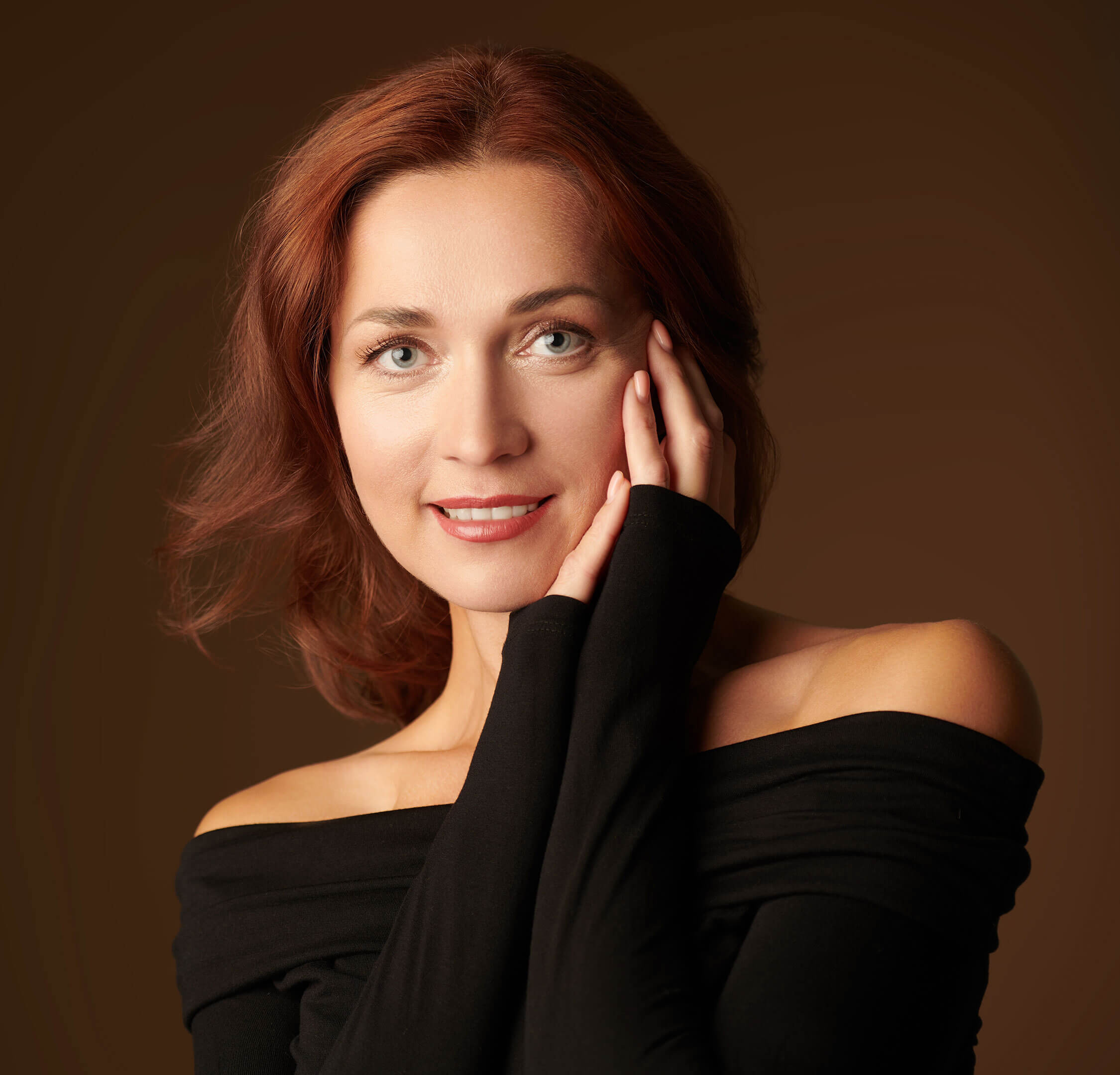 Red headed woman with her left hand on her left cheek in a black off the shoulder blouse